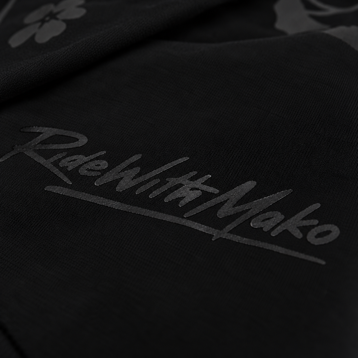 Close-up of a black fabric product featuring a tonal dark gray 'RideWithMako' script logo and a subtle floral pattern.